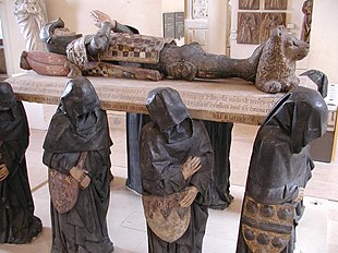 A large sculpture of six life-sized black-cloaked men, their faces obscured by their hoods, carrying a slab upon which lies the supine effigy of a knight, with hands folded together in prayer. His head rests on a pillow, and his feet on a small reclining lion.
