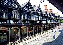 A street containing a range of two-storey shops on the left seen from a slightly elevated position. The lower storey contains modern shop fronts behind an arcade; the upper storey is timber-framed and contains a variety of windows and decorated gables.