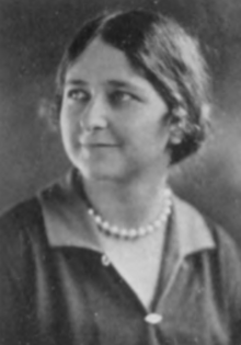 A young white woman wearing a dark blouse with a collar, and a strand of beads at her neck