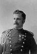 Head and shoulders of a white man with wavy hair and a mustache, wearing a double-breasted military jacket with shoulder cords and two medals hanging from ribbons on the left breast.