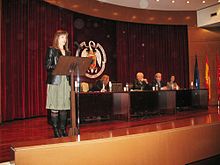 Woman speaking onstage, with others seated at a dais