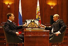 Medvedev and Putin, both white men, wearing suits sitting at a wodden desk across from one another. There are flags in the background.