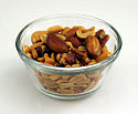 A photograph of a small, clear bowl of mixed nuts displaying large nuts on top and peanuts below, all on a white surface in front of a white background