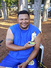 A man wearing a blue sleeveless T-shirt and blue shorts smiles while holding a bobblehead depicting himself in a white baseball uniform.