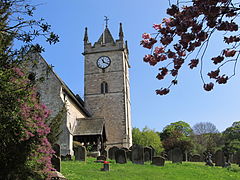 A bright spring day on a country churchyard, surrounded by mature trees. The church nave is on the extreme left, and at the far end is tall, square, tower capped by a pointed wooden roof, not quite a spire, and with pinacles on all 4 corners. A bold white clockface is on the tower. The church has an ope porch, with pillars of wood. Dark, old, gravestones are scattered in the green grass.