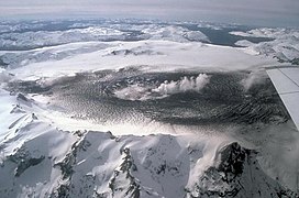 A steaming depression, surrounded by dark ash deposits, in the middle of a circular ice field