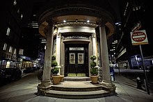 The entrance at Beaver and South William Streets at night. Two steps lead up to a double doorway with lights. There are columns on either side of the doorway.
