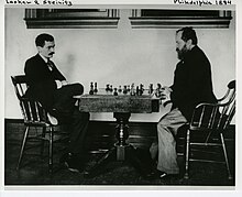Emanuel Lasker and Wilhelm Steinitz both sitting down at a chessboard during a game. Steinitz has the White pieces, and Lasker has the Black pieces.