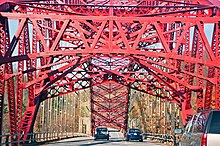 A red steel bridge arch is seen from a car going under the metalwork along the bridge deck