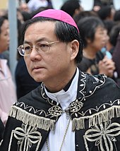 A bespectacled Chinese man wearing a black cope with silver pattern and trimming.