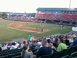 A baseball game being played on a green field surrounded by a grandstand with red and green seats