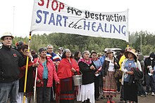 A group of people gather outdoors with a large banner reading "Stop the destruction, start the healing".