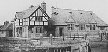 An old photograph showing, from a slight angle, a house on the left and a school on the right. The house is in stone with a jettied timber-framed upper storey; the school is in stone with three gables in a steep roof containing three dormers and, to the left a bell cote.