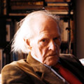 Photograph of an unsmiling, elderly man looking side on into camera. His hair is white and collar length. He wears a brown jacket, blue-collared shirt and red tie. He appears to be seated in front of a bookcase.