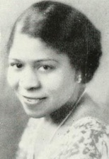 A smiling African-American woman with short hair, wearing a print dress and pearls.