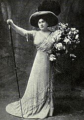 Woman standing in a dramatic pose, right arm raised, left arm holding a large bouquet. She is wearing a long formal gown and a wide-brimmed hat.