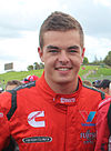 A teenaged man wearing red racing overalls with sponsors logos is smiling at the camera