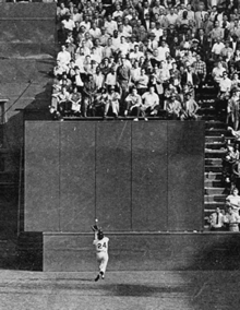 Nearly at a wall, Mays reaches up underhanded to catch a ball falling towards him while over 30 feet above, several rows of fans stare down at him