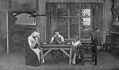 Indoor scene with a woman, a man and a young girl, seated at a cottage table, heads bowed for grace