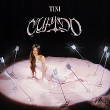 Tini sitting in the middle of a pink circle, surrounded by arrows lodged in the ground. The arrows have heart shaped tips. The background is black and her name Tini and the album title, which is printed in a pink swirling font, appear in capital letters above.
