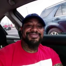 A man smiling in his car at a parking lot.