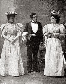 Butler between two young women, all dark-haired and all standing, in a garden setting; the women, both in hats, regard each other angrily