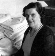 Black and white portrait photograph of MacKinnon, looking into the camera and smiling. There is hospital linens behind her.