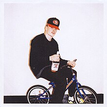 A photo of the artist sat on a small bicycle and holding a bottle of alcohol in one hand.