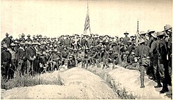 A black-and-white photo of US Army soldiers on 3 July 1898, in an unside down V type formation on top of Kettle Hill, two American flags in center and right. Soldiers facing camera.