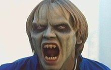 A blond man with greenish skin, blond hair and a blue shirt looks off-screen, opening his mouth wide to reveal sharp pointed teeth.