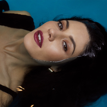 A color photograph of Marina Diamandis laying down in water with her face exposed.