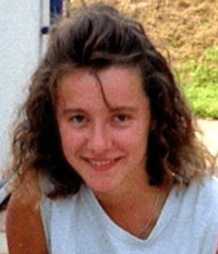 A Caucasian female, probably in her late teens, with light brown hair and brown eyes wearing a white sleeveless shirt looking at the camera and smiling. Visible in the background are a red-and-white patio, a lawn and a white-painted building with an attached black drainpipe.