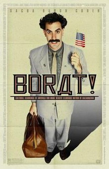 Sacha Baron Cohen as Borat wearing a business suit while holding a suitcase on his right hand and an American flag on his left hand.