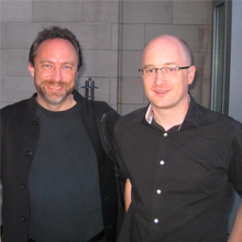 A photo of two men wearing black shirts standing outside a concrete building