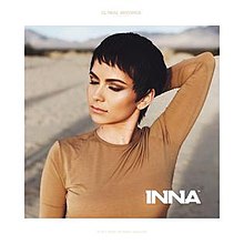 A shot of a short-haired Inna posing in front of a desert, wearing a beige bluse and touching her head with her right hand.