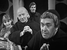 The Monk (right) looks eagerly to the left of the camera. In the background is the Doctor (dressed in monk clothing), Vicki, and Steven. They are standing in the Monk's TARDIS at the main console; the roundel walls can be seen in the far background, and the doors are open.