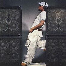 An image of Musiq standing between loudspeakers stacked on top of each other, sporting a white t-shirt, jeans, hat and black headphones.