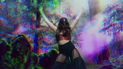 Dua Lipa with her hands up, wearing a black bra and black mesh pants, dancing in a technicoloured forest.