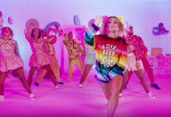 Shot of Delia wearing a multicolored sequined anorak while background dancers are shown in a background decorated with candy.