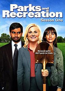 The DVD cover shows three people standing side-by-side. In the middle, a blond woman wears a gray dress suit, smiling and holding a golden shovel. On the right, a brown-haired woman in a green jacket looks at her and smiles. On the left, a black-haired man with a beard, wearing a gray suit and green tie, looks at the middle figure while smirking. Above the trio is the text, "Parks and Recreation Season One".