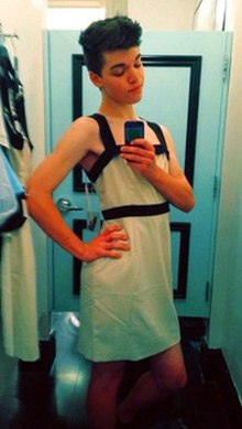 Teenage, white, trans girl with dark hair wearing a white dress and posing in front of a mirror, taking a photograph of herself using a camera phone