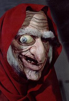 A latex mask of a wrinkled old woman with long white hair, bushy eyebrows, a large nose, and a bulging right eye wearing a red hooded cloak