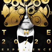 Black and white photo of a man, wearing a suit and bowtie, facing forward - the upper half of his head is covered by a gold phoropter - the background is entirely black. The lower half of the image features golden, shiny text reading "THE 20/20 EXPERIENCE".