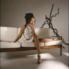 Leigh-Anne sitting on a white sofa in a shadowy white room, with a wooden sculpture behind her