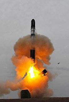 A rocket climbing out of a hole in the ground through an orange cloud of dust and smoke, with a metal ring falling away from the bottom of the rocket