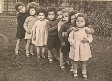 8 mixed race children standing in a row in a garden in the 1940s at Holnicote House in England. The children are wearing dresses or shorts.