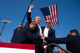 A man with his fist held above his head with blood on his ear and cheek. An American flag flutters in the air behind him. Two federal agents shield and escort him.