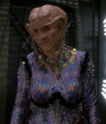 A white woman in the heavy orange makeup and prostheses typical of Ferengi looks to the camera's left; she is wearing a garish, skintight outfit and excessive jewelry from her clothes, neck, and lobes