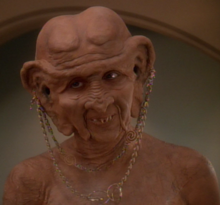 A white woman in the heavy orange makeup and prostheses typical of Ferengi looks to the camera's left; she is nude, baring her upper chest and shoulders