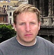 A young Caucasian man with close-cropped blond hair wearing a gray shirt and looking at the camera, with some older, classically styled buildings behind him